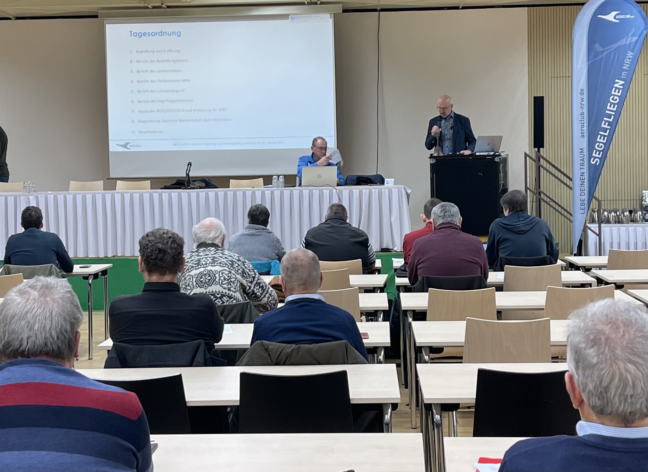 IMG_6797 Jahreshauptversammlung der Sportfachgruppe Segelflug und Motorsegler 2023. Mehrere Teilnehmer sitzen vor einer Bühne an tischen, die vorher in reihen aufgestellt wurden und hören aufmerksam der Präsentation zu.