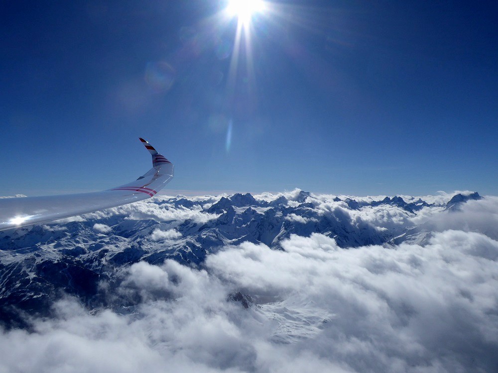 Luftaufnahme aus einem Segelflieger raus. Man sieht einen klaren blauen Himmel mit Sonnenschein. Darunter befinden sich eingeschneite Berg Gipfel in Wolken gehüllt.