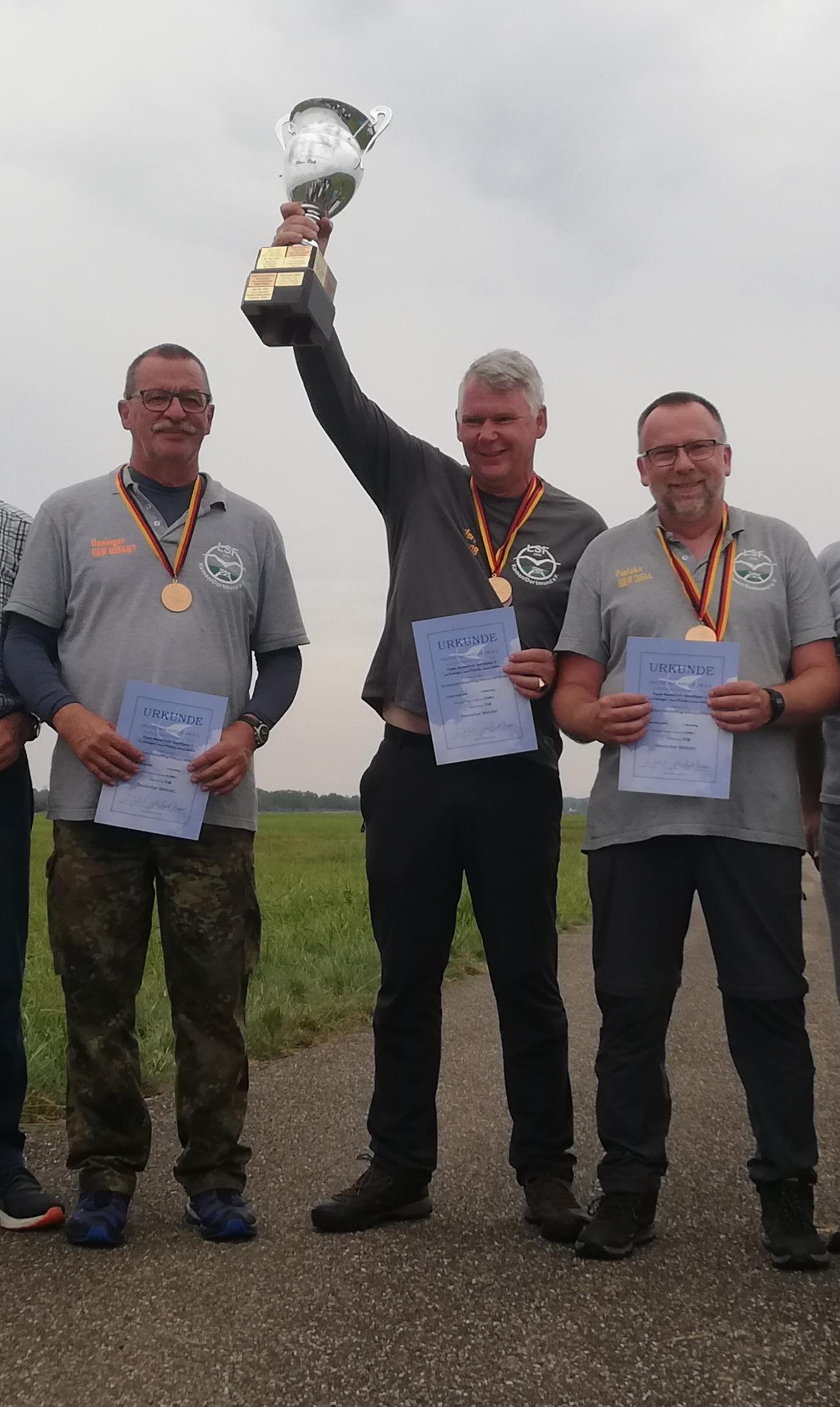 Team F1B Team F1B mit Urkunden und Medaillen die sie in die Kamera präsentieren. Ein Mann in der Mitte hält einen silbernen Pokal in die Luft. Im Hintergrund sieht man ein weites, grünes Feld und einen grauen Himmel.