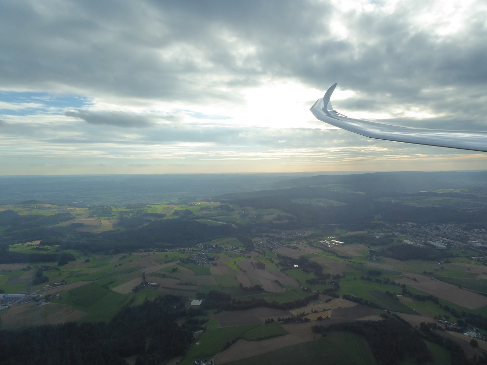 Abendstimmung über den Mittelgebirgen, bei einem Segelflug. Der Himmel ist grau Bewölkt mit ein paar wenigen Sonnenstrahlen. Unter dem Flieger befinden sich weite Felder und ein Dorf.