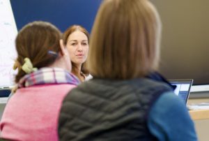 Eine Frau mit mittel langen Haaren, sitzt an einem Tisch und schaut in die Kamera. Es handelt sich um Anne Hennig, die einen Workshop gibt. Vor ihr sitzen zwei Frauen mit einem Laptop, mit dem Rücken zur Kamera.