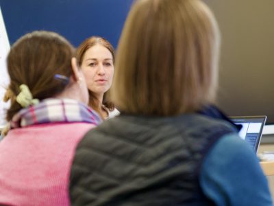 MA306365-scaled-400×300 Eine Frau mit mittel langen Haaren, sitzt an einem Tisch und schaut in die Kamera. Es handelt sich um Anne Hennig, die einen Workshop gibt. Vor ihr sitzen zwei Frauen mit einem Laptop, mit dem Rücken zur Kamera.