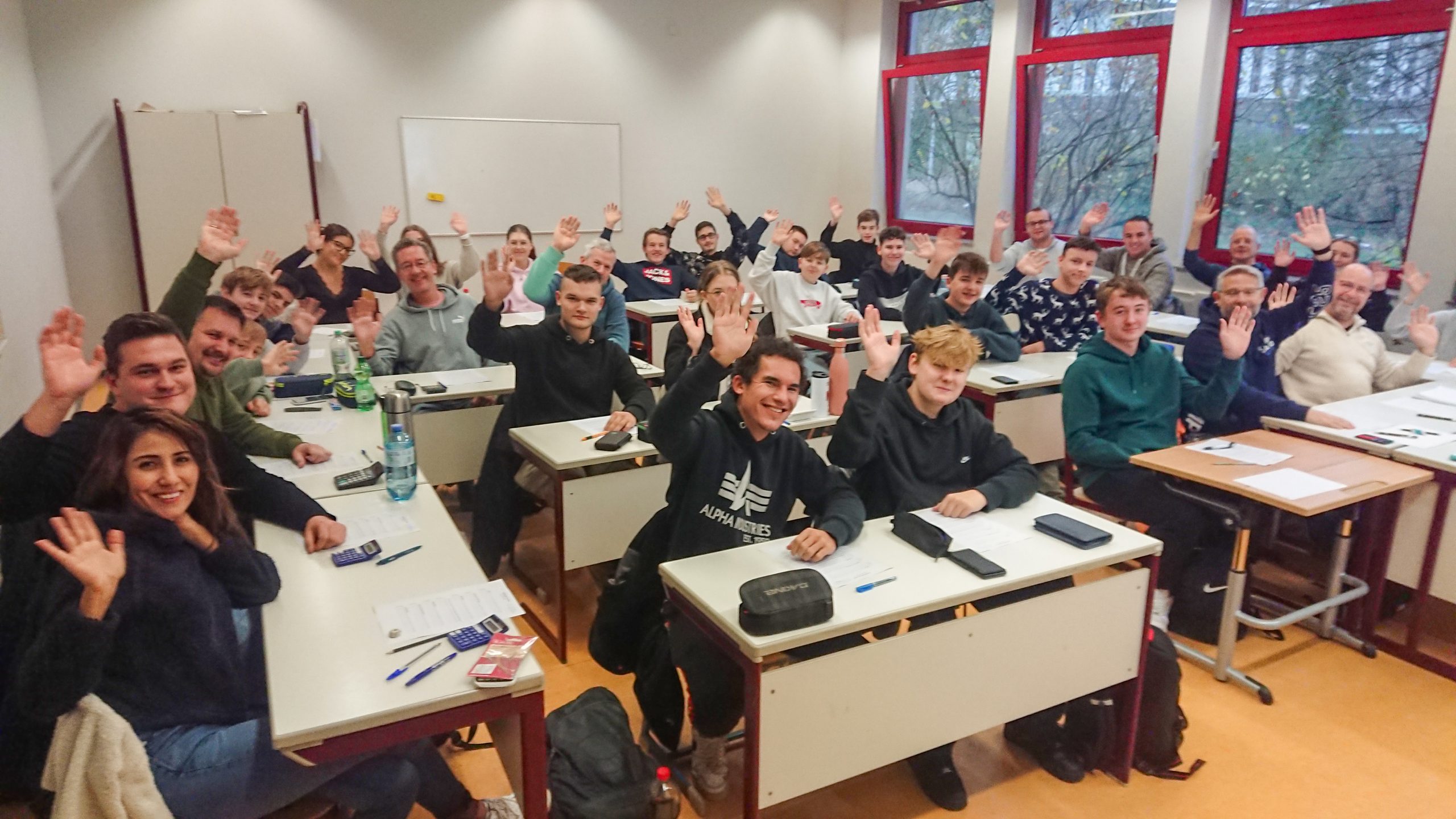 Kursfoto GK 2024 Teilnehmende des 50. Theoriekurses im November und Dezember 2024. Die Teilnehmenden sitzen in einem Klassenraum, an Tischreihen nebeneinander und Winken in die Kamera.