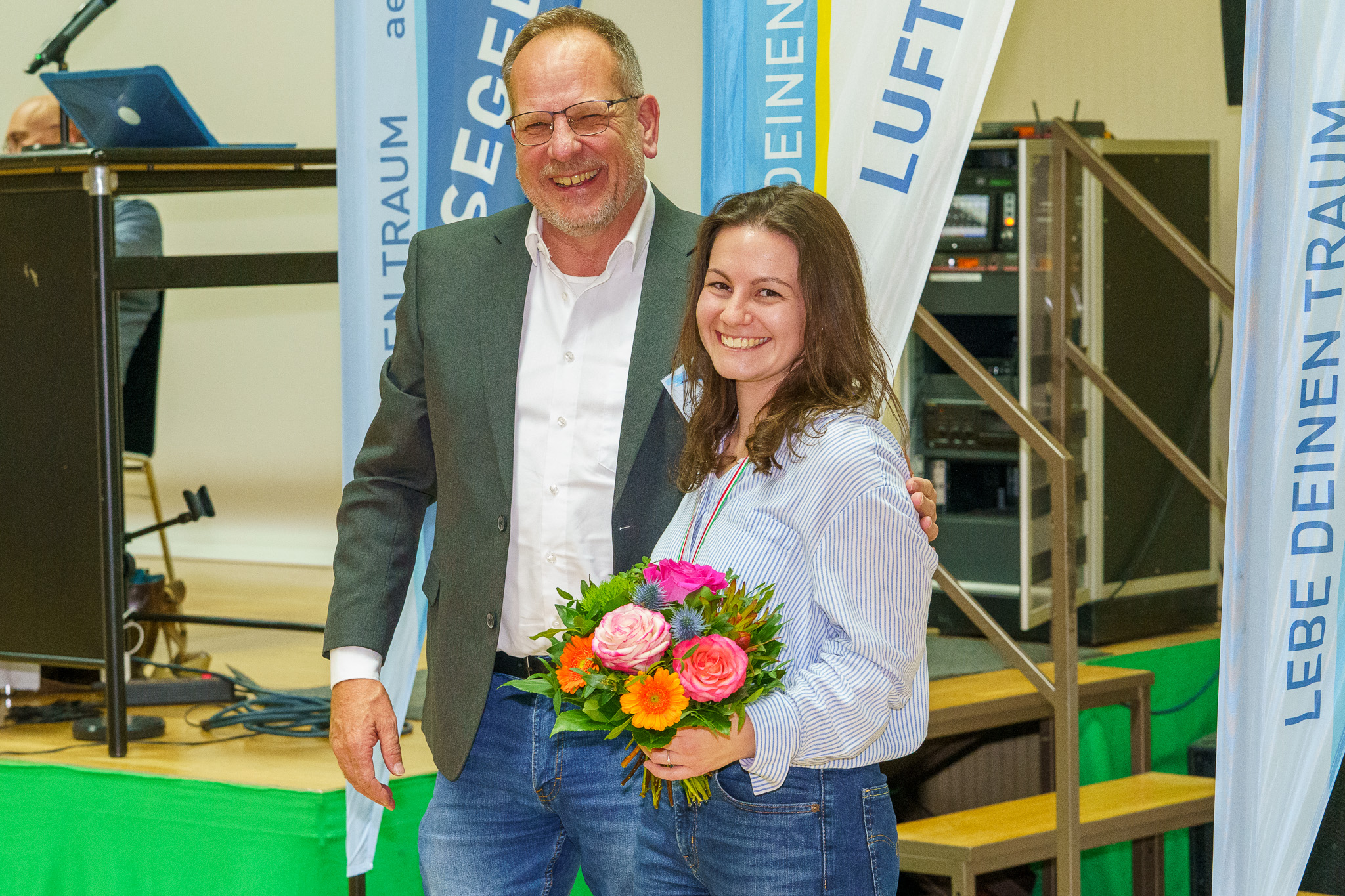 MA306443 Laura Budny und Aeroclub-Präsident Volker Engelmann stehen Arm in Arm vor einem Aeroclub Banner. Sie hält einen bunten Strauß Blumen in der Hand und beide lächeln in die Kamera. Sie wird mit der Plakette des Landesverbandes geehrt.