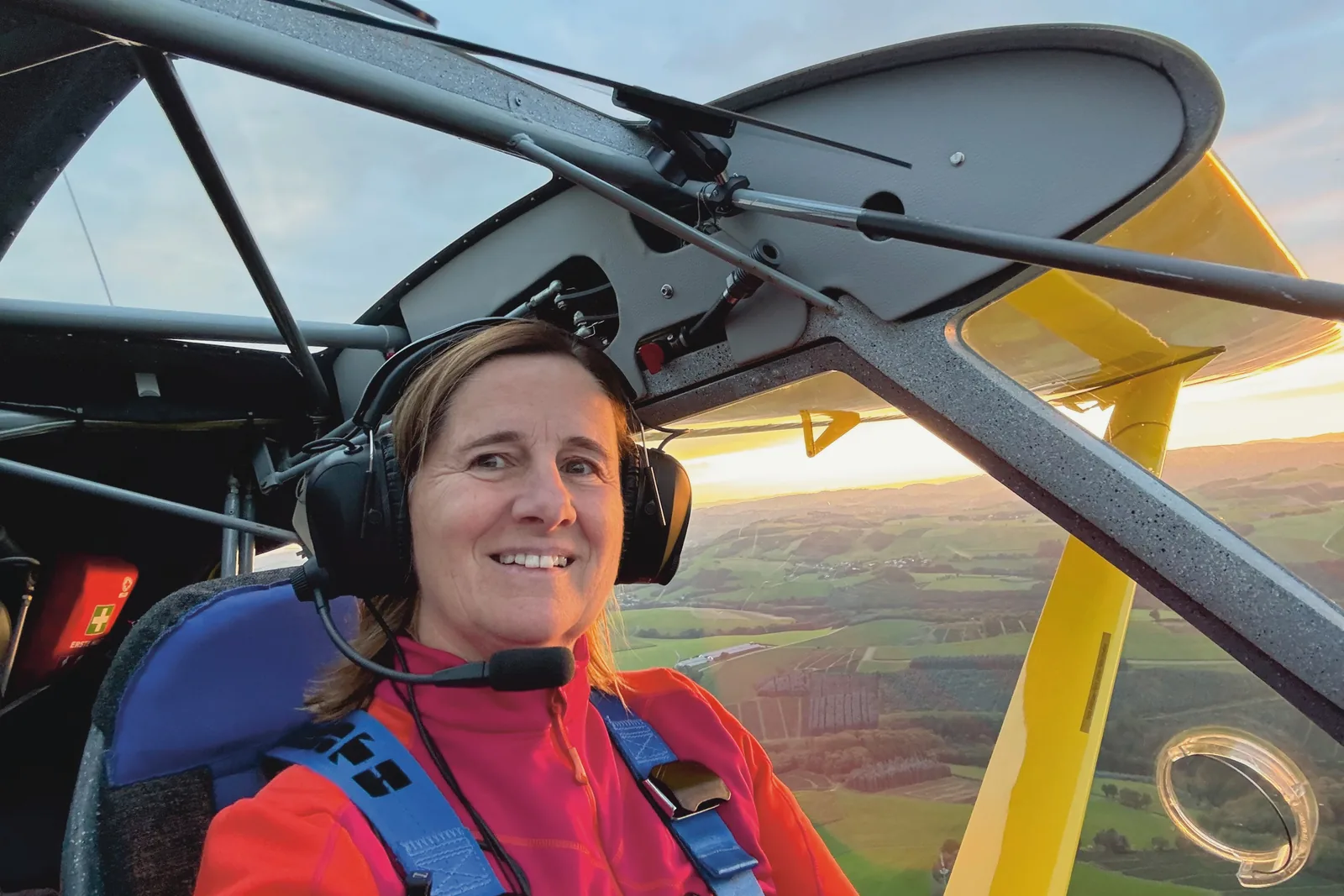 Pilotin mit Headset im Cockpit eines Hochdeckers, im Hintergrund Abendsonne über einer Hügellandschaft
