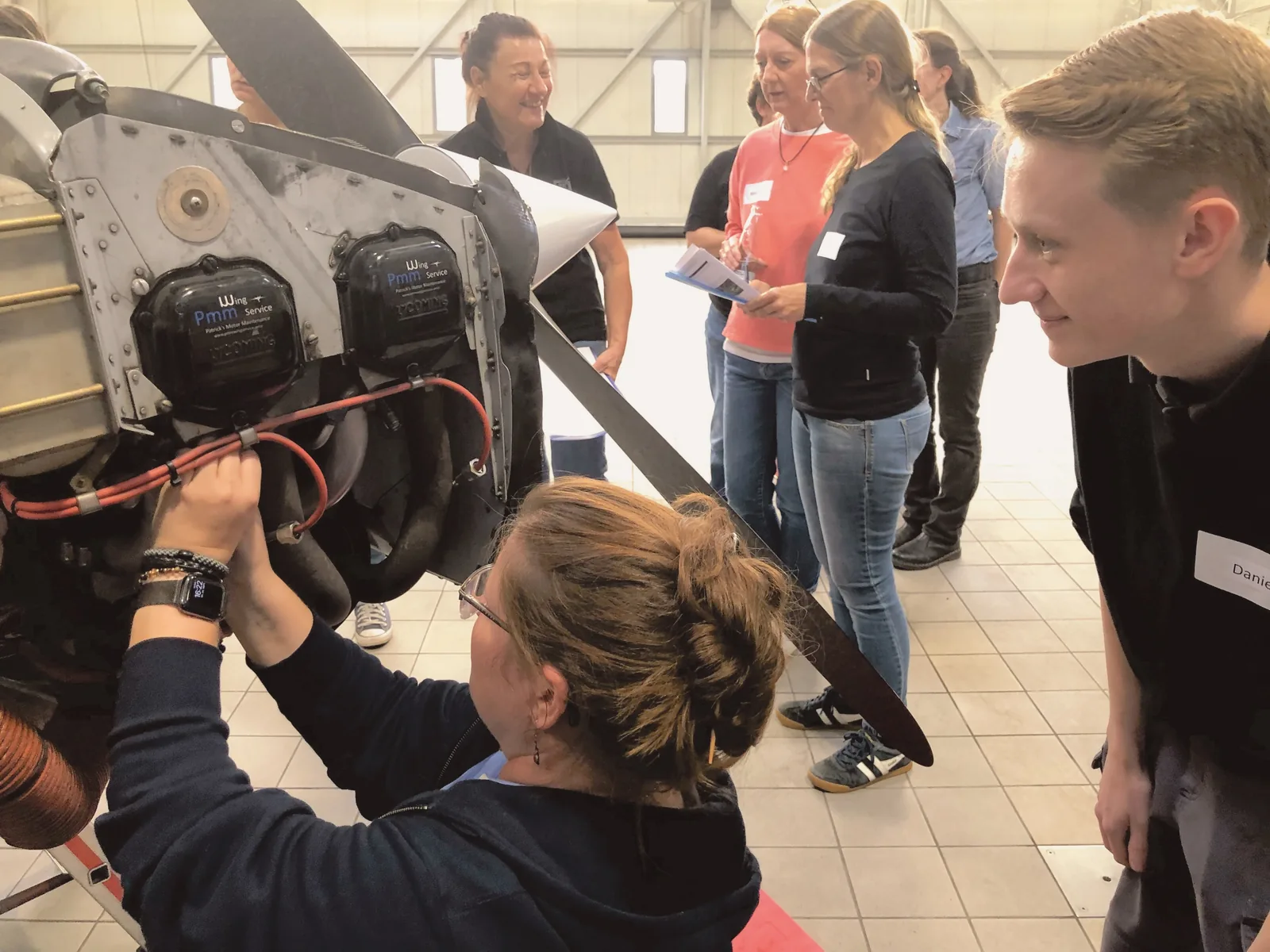 Pilotin arbeitet an einem offenen Flugzeugmotor, weitere Teilnehmende schauen zu