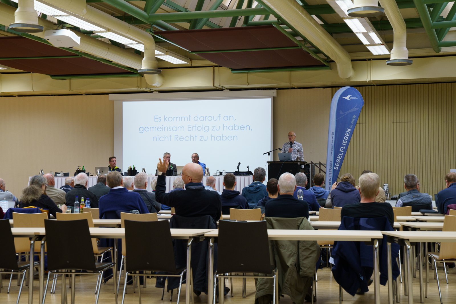 Vereinsvertreterinnen und Vereinsvertreter bei der Jahreshauptversammlung der Segelflieger, vorn Rednerpult, Podium und Banner Segelfliegen in NRW