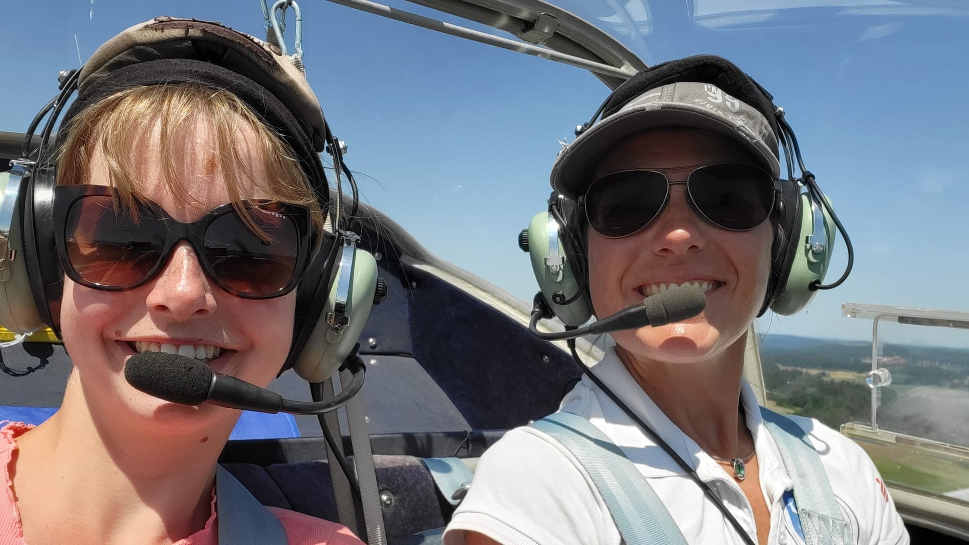 Zwei Pilotinnen mit Headsets und Sonnenbrillen lachen im Cockpit eines Kleinflugzeugs in die Kamera