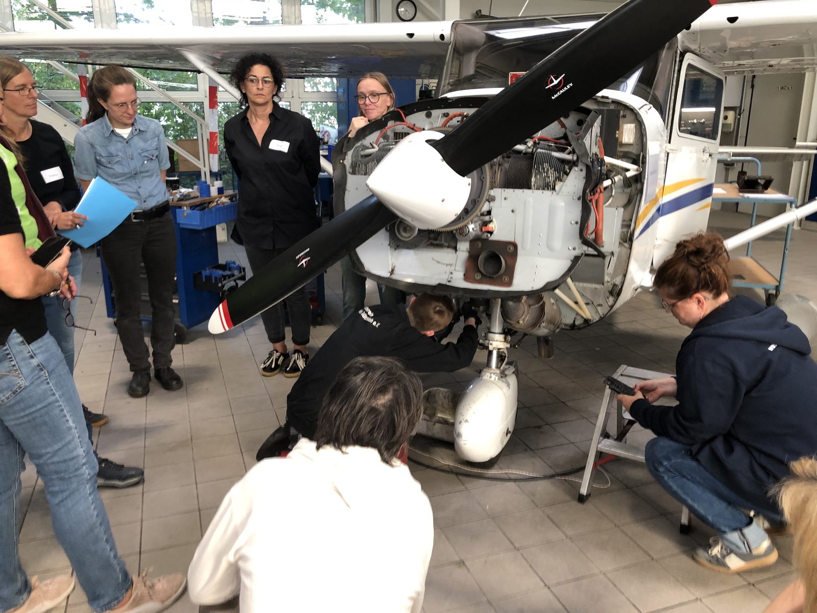 Teilnehmerinnen des Workshops an der geöffneten Motorverkleidung einer Cessna 172 R im Hangar